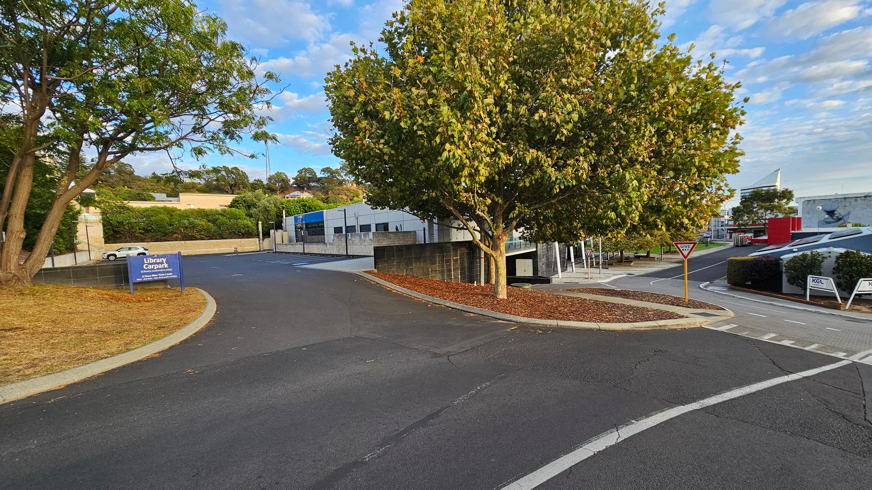 A road curves around a large tree in the centre, with a "Give Way" sign nearby. On the left, a sign indicates a "Library Carpark" with a white car parked in the distance. The background features a building with a blue stripe and more trees. On the right, there are additional buildings, including one with a red section, and signs marked "KGL". The ground is paved, and the area around the tree is covered with mulch.
