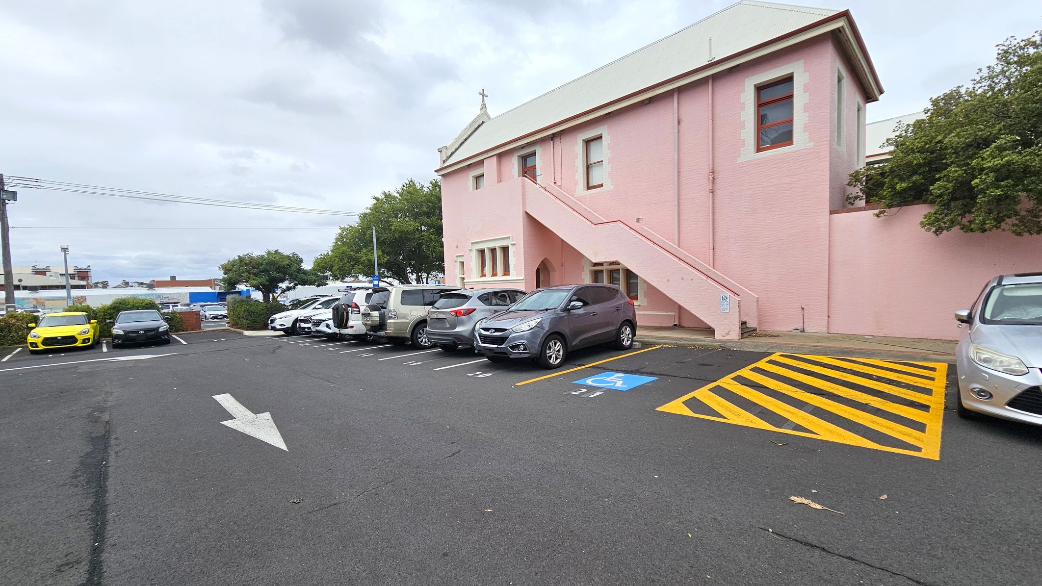 A car park with several vehicles lined up on the left, including a yellow car and a black car. A pink building with a staircase is on the right, featuring windows and a small cross on the roof. A designated disabled parking space with blue and yellow markings is visible in the foreground. Trees and other buildings are in the background, and an arrow is painted on the asphalt, indicating traffic direction.