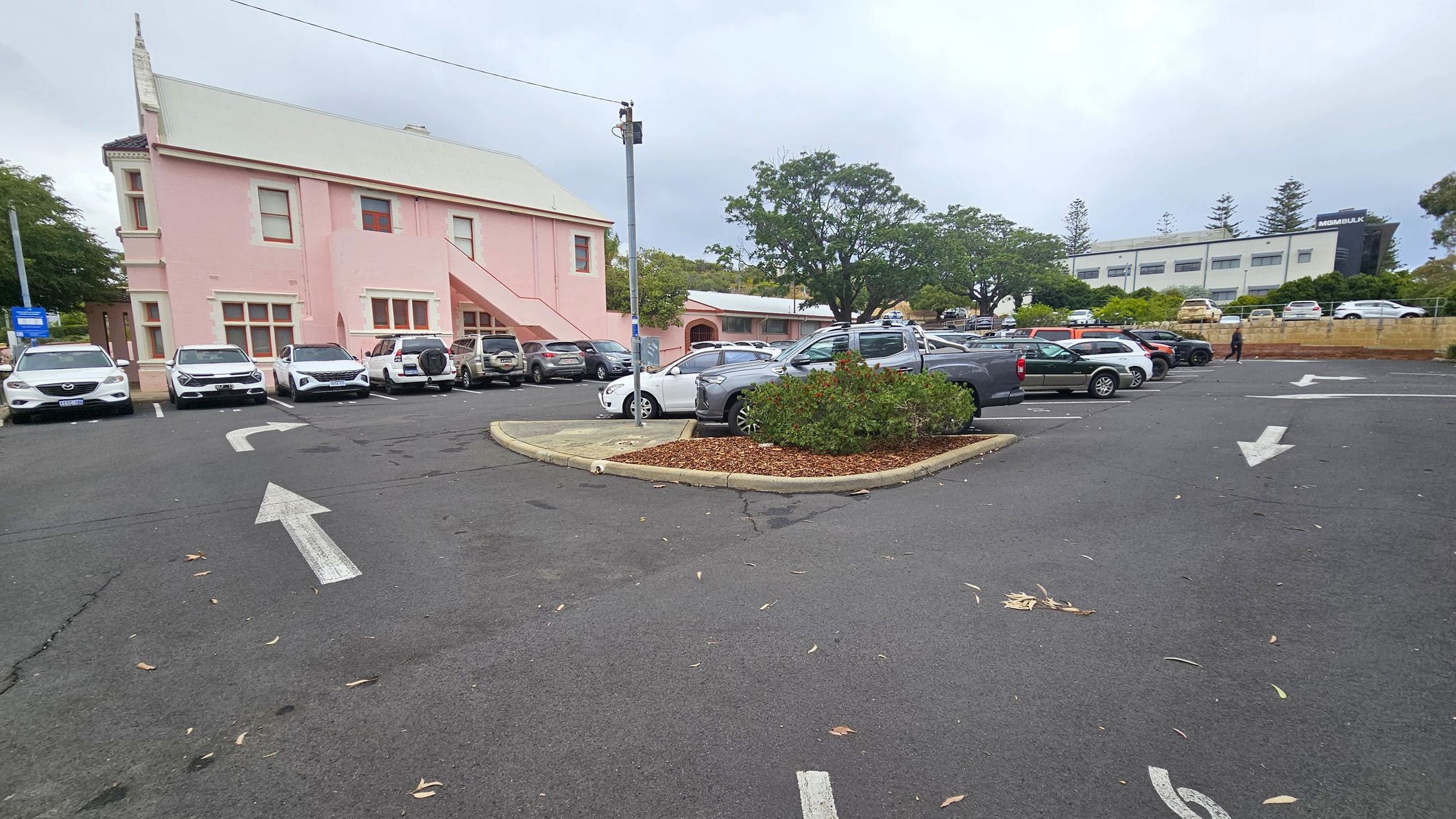 A car park with several vehicles is visible. On the left, there's a pink building with a sloped roof and external staircase. In the centre, a small landscaped area with shrubs is surrounded by parked cars. On the right, more cars are parked, and a person is walking. In the background, a white building with the word "MSMBULK" is partially visible, along with trees and a low wall. The asphalt surface has directional arrows painted on it.