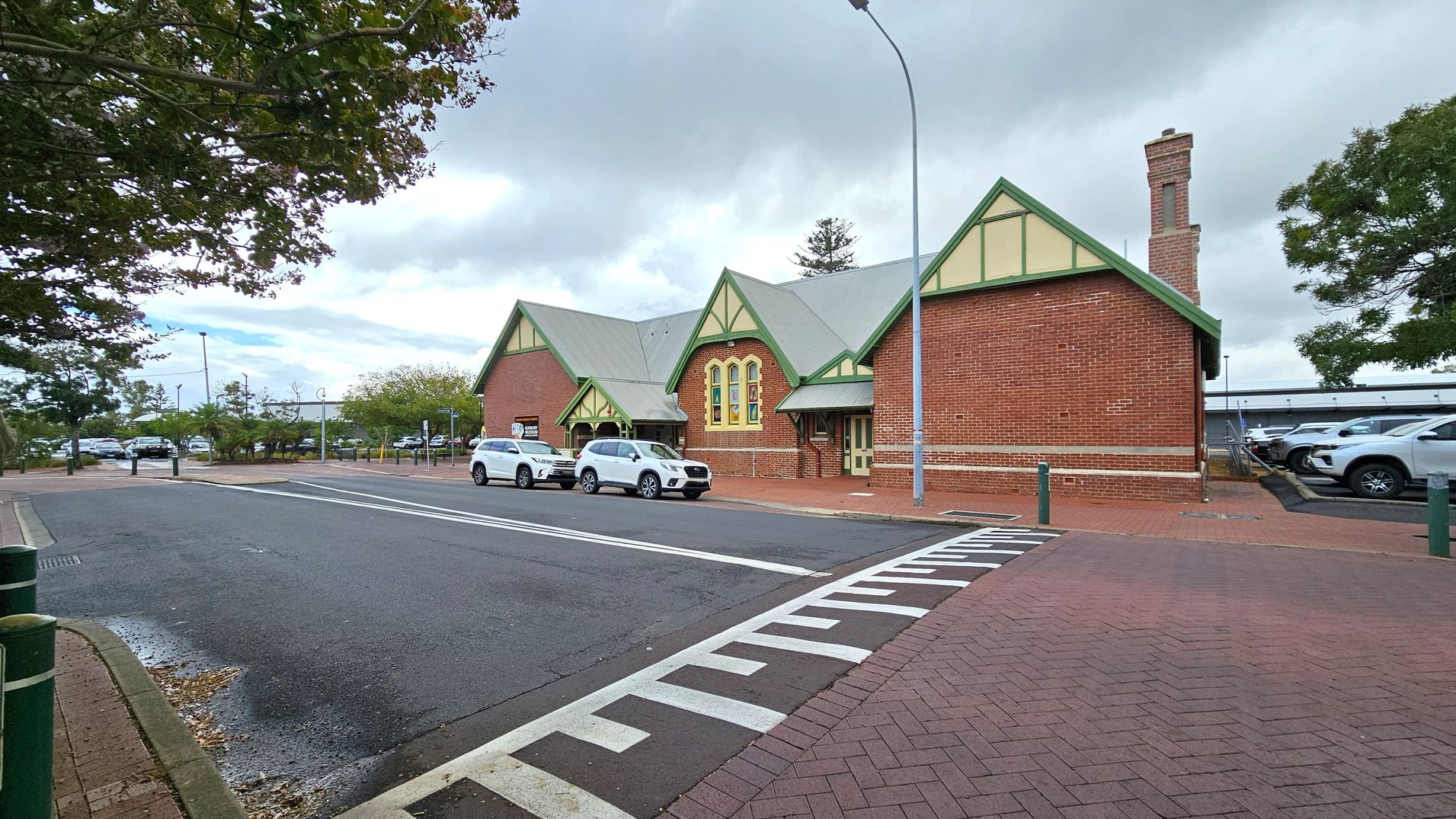 A red brick building with green trim and a gabled roof is on the right. It has a chimney and several windows. Two white cars are parked in front. The road in the foreground has a pedestrian crossing with white markings. On the left, there are trees and a parking area with more cars. A tall streetlamp is near the building. The pavement is red brick, and the sky is overcast.