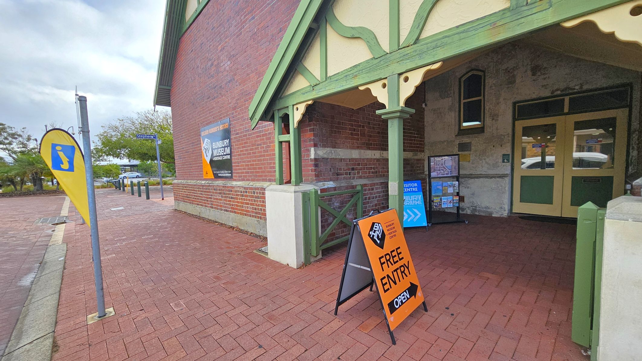 A brick building with green trim and a gabled roof on the right. The entrance features double doors and a signboard with maps and brochures. An orange sign on the pavement reads "Free Entry" and "Open." A yellow flag with an information symbol is on the left, near a street sign for "Stephen St." The pavement is red brick, and there are trees and a parked car in the background.