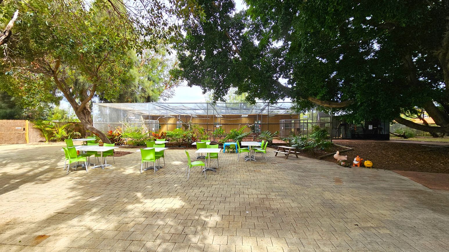 A paved outdoor area with several tables and bright green chairs scattered around. On the left, a large tree provides shade, and there are plants and a wooden fence in the background. In the centre, a structure with a mesh roof is visible, possibly an aviary or greenhouse. On the right, a picnic bench and some decorative items, including a pumpkin, are placed near the base of another large tree. The ground is covered with light-coloured paving stones.