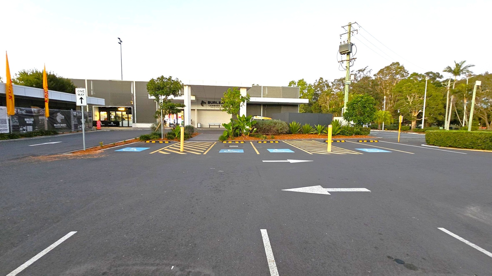 A car park with marked spaces, including two for disabled parking, is in the foreground. A white arrow on the asphalt indicates a one-way direction. In the background, a building with a sign and glass entrance is visible, surrounded by trees and greenery. A "One Way" sign is on the left, and a power pole with cables is on the right. The area is bordered by hedges and trees.