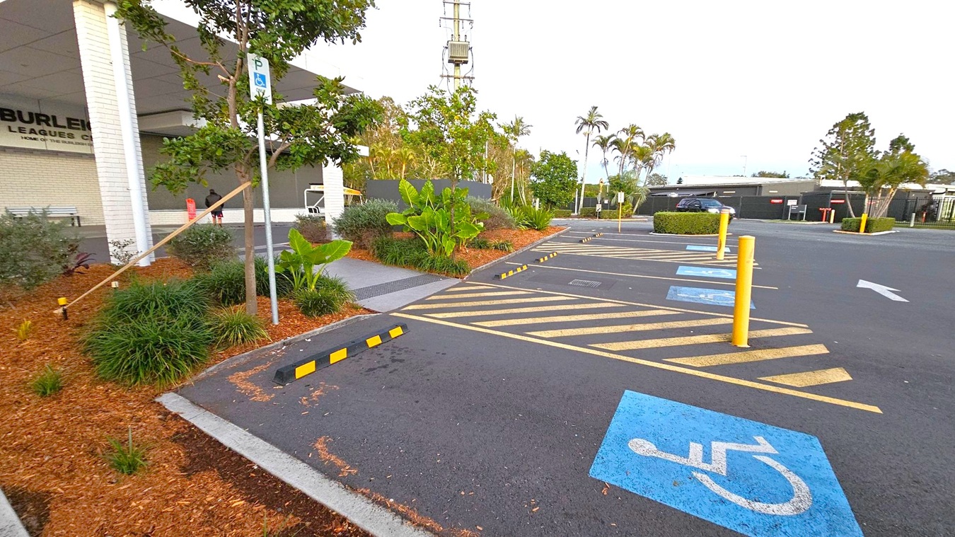 A parking area with designated disabled parking spaces marked in blue on the right. Yellow-striped zones and bollards separate the spaces. On the left, a landscaped area with mulch, small plants, and a tree next to a white brick building. A signpost indicates parking for disabled individuals. In the background, there are more trees, a tall structure, and a black building. The ground is asphalt, and the area is well-maintained.