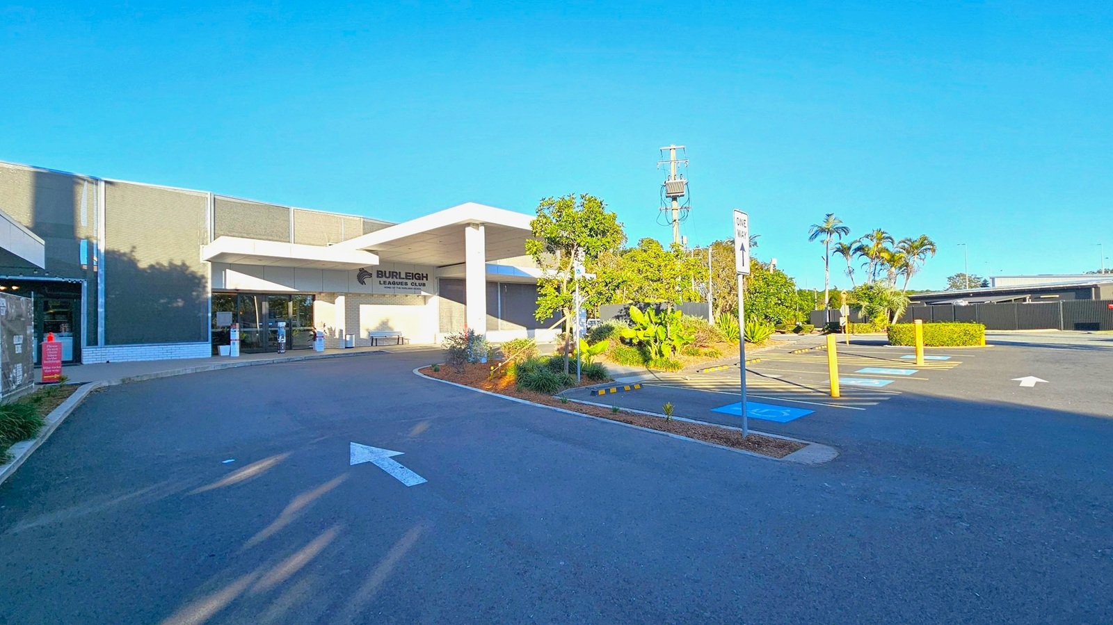 A building with a sign reading "Burleigh Leagues Club" is on the left, featuring a modern design with large windows and a flat roof. In front, there are landscaped areas with small trees and shrubs. The car park on the right has marked spaces, including accessible parking, and a "One Way" sign. The ground is paved, and there are yellow bollards near the parking spaces. In the background, there are more trees and a power pole.