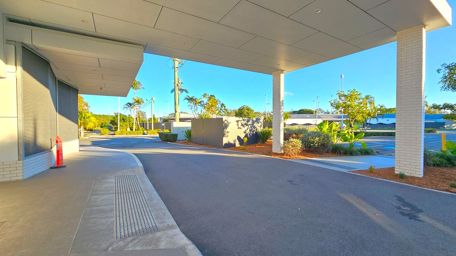 A covered walkway with a tiled ceiling and a white brick pillar on the right. The left side features a building with closed metal shutters and a red fire hydrant. The ground is a mix of concrete and asphalt. In the background, there are palm trees, shrubs, and a parking area with a few parked cars. The sky is clear and blue.