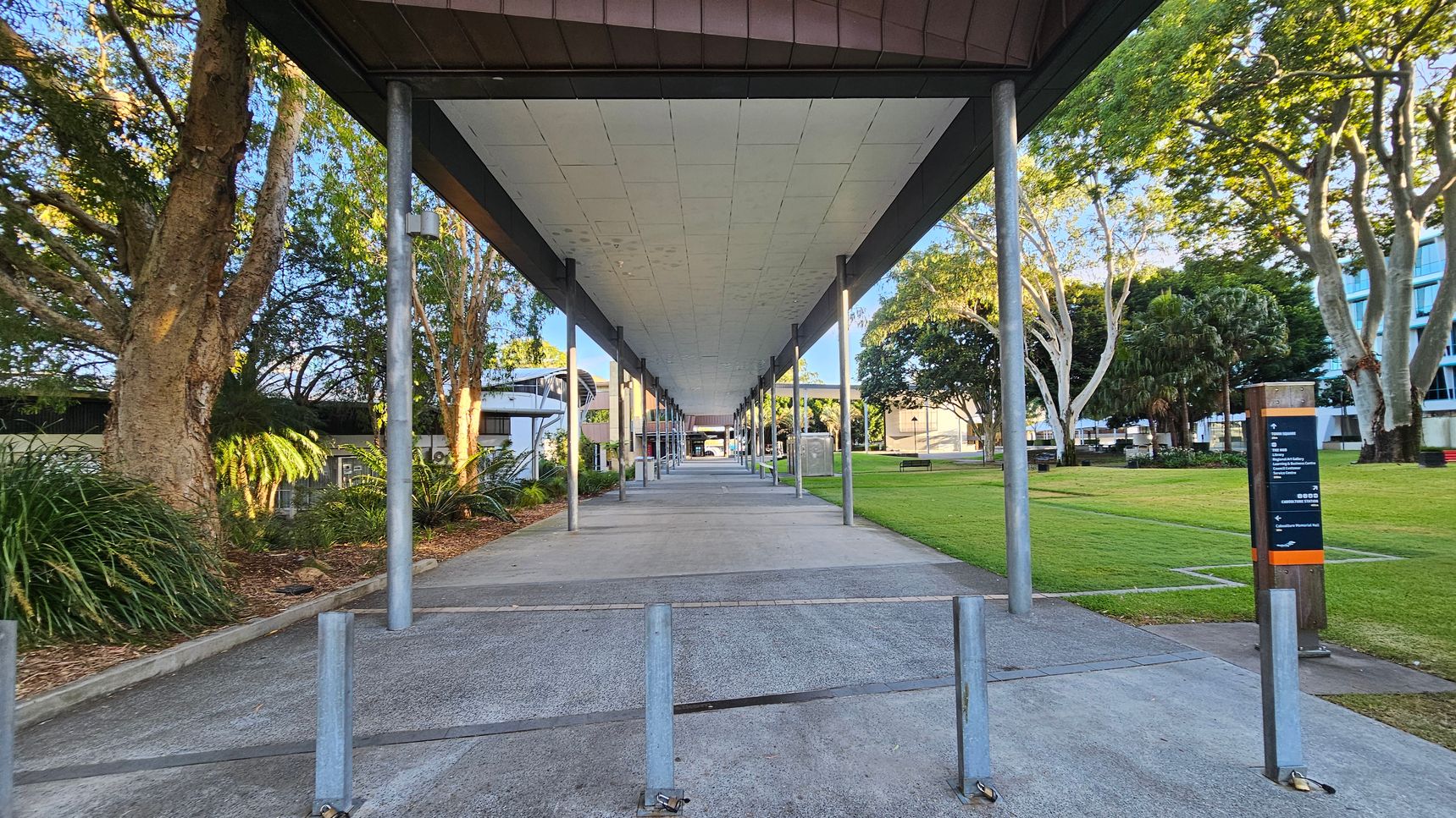 A covered walkway with a series of metal poles runs through the centre, leading to a distant building. On the left, there are large trees and dense greenery. On the right, a grassy area with more trees and a signpost. The ceiling of the walkway is flat and light-coloured, while the ground is paved with concrete.