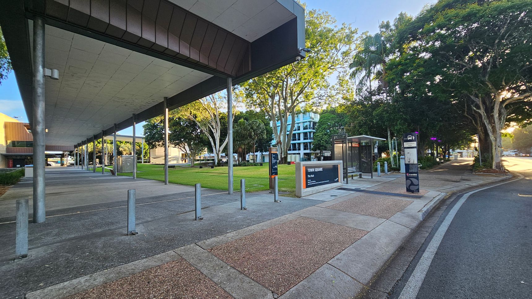 A modern urban scene with a covered walkway on the left, supported by metal poles. The ground is a mix of concrete and textured paving. In the centre, there's a grassy area with trees and a sign reading "Town Square." A bus stop with a shelter and signage is on the right, next to a road curving into the distance. Trees with dense foliage line the right side, and a multi-storey building is visible in the background. The lighting suggests early morning or late afternoon.