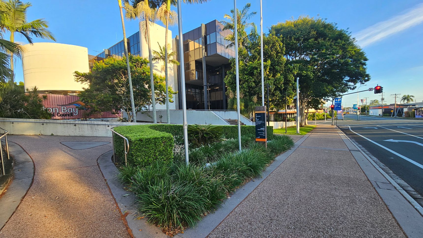 A modern building with reflective glass panels is in the centre, surrounded by tall palm trees and lush greenery. On the left, a curved pathway leads up to the entrance, bordered by manicured hedges. A sign with partial text is visible. On the right, a wide pavement runs alongside a road with marked lanes and a traffic light. The area is well-maintained, with a mix of natural and urban elements.