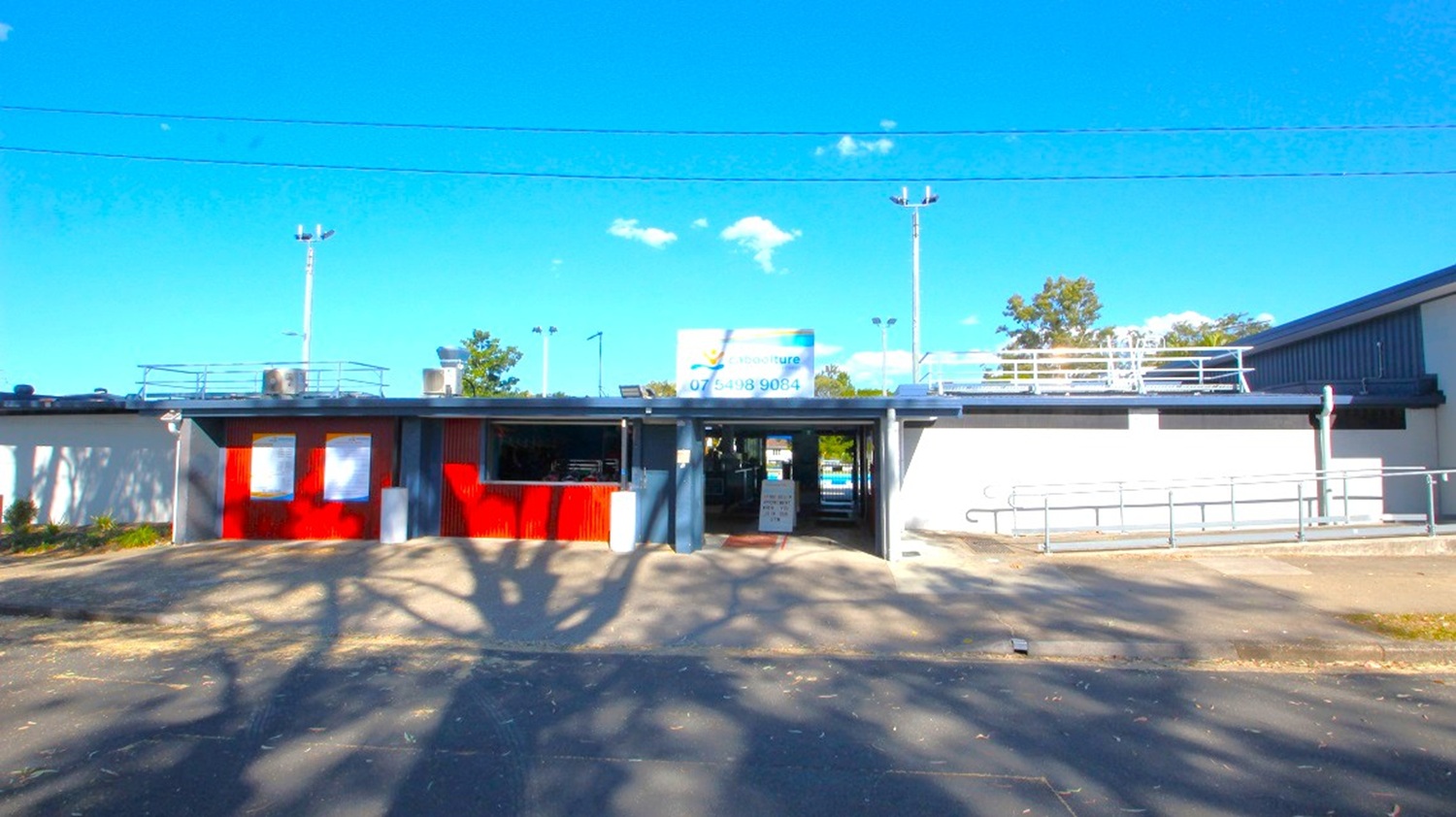 A single-storey building with a flat roof and a sign on top. The left side features red panels and a small window, while the right side has a grey wall with a ramp and railings. In the centre, there's an entrance with a signboard on the ground. The foreground shows a paved area with tree shadows. Floodlights and trees are visible in the background.