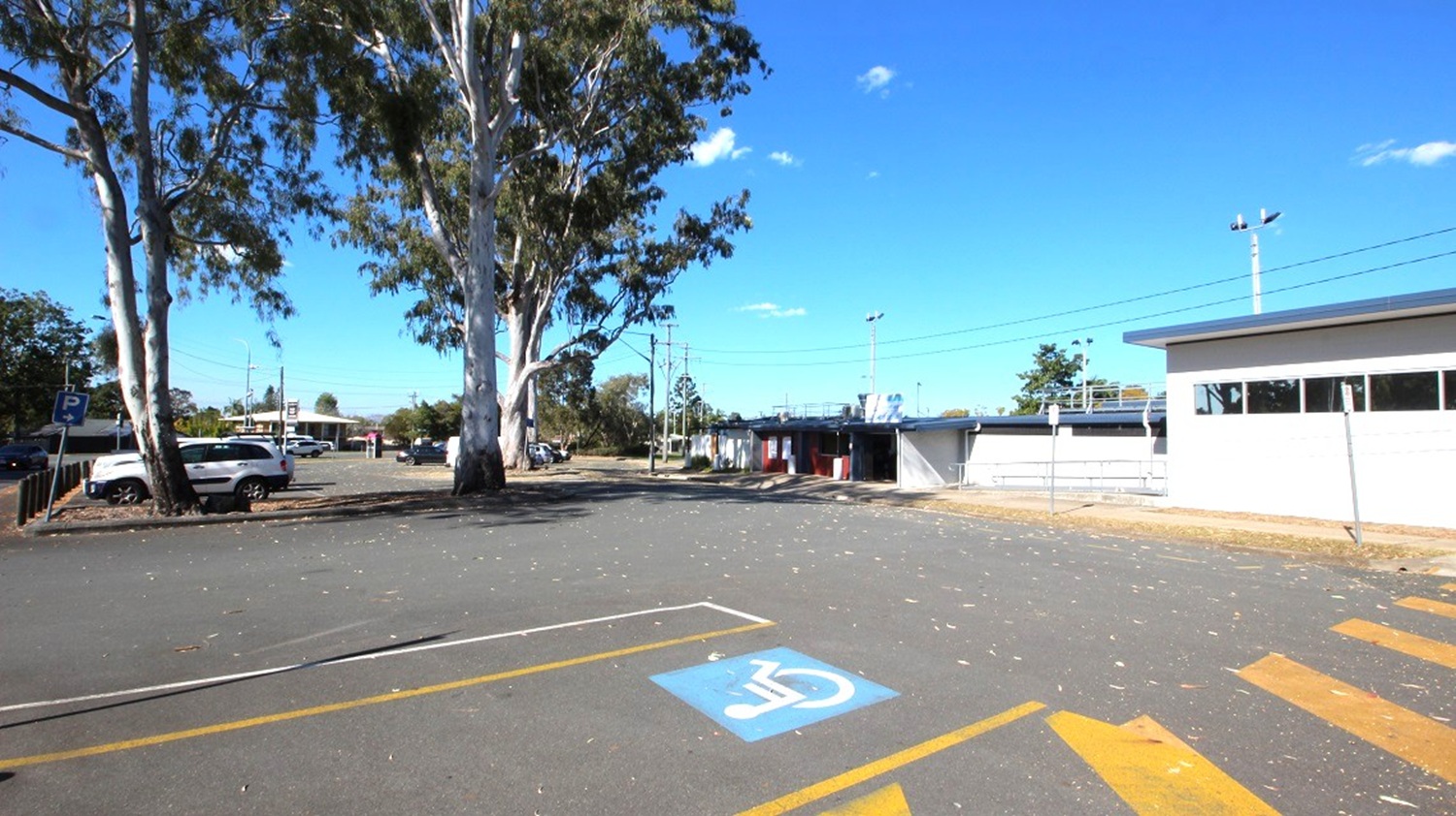 A parking area with a marked disabled parking space in the foreground. On the left, there are several large trees and a few parked vehicles. To the right, a low building with a flat roof and windows. In the background, more buildings and a clear blue sky. The ground is paved, with some scattered leaves.