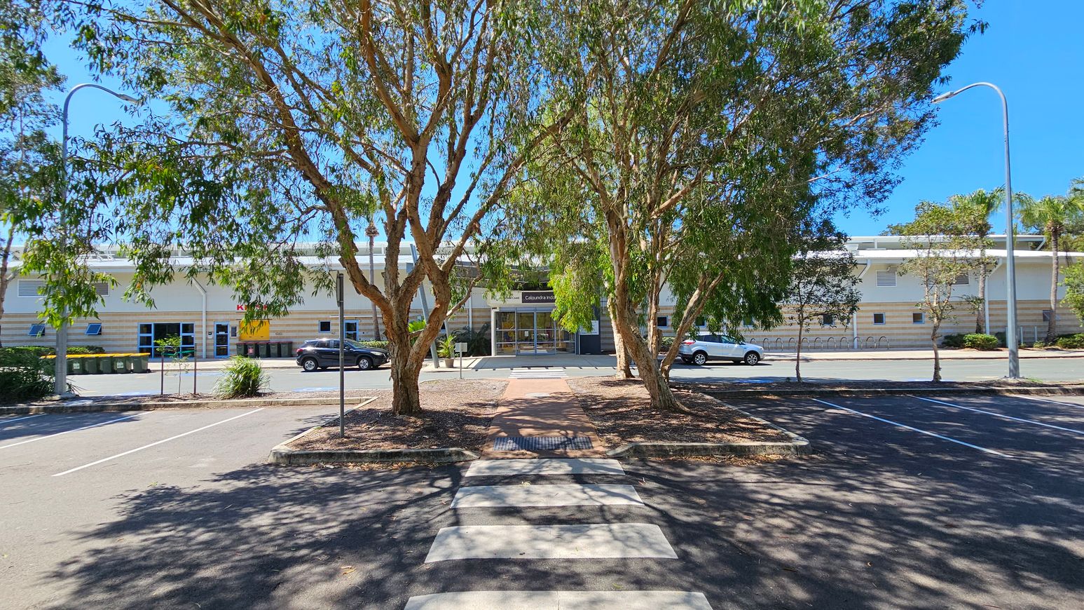 A car park with a zebra crossing in the foreground leads to a building in the background. The building has a modern design with a glass entrance and a sign above the door. Two large trees with green foliage are in the centre, casting shadows on the ground. Cars are parked along the street in front of the building. Street lamps are visible on both sides, and there are several bins to the left. The sky is clear and blue.