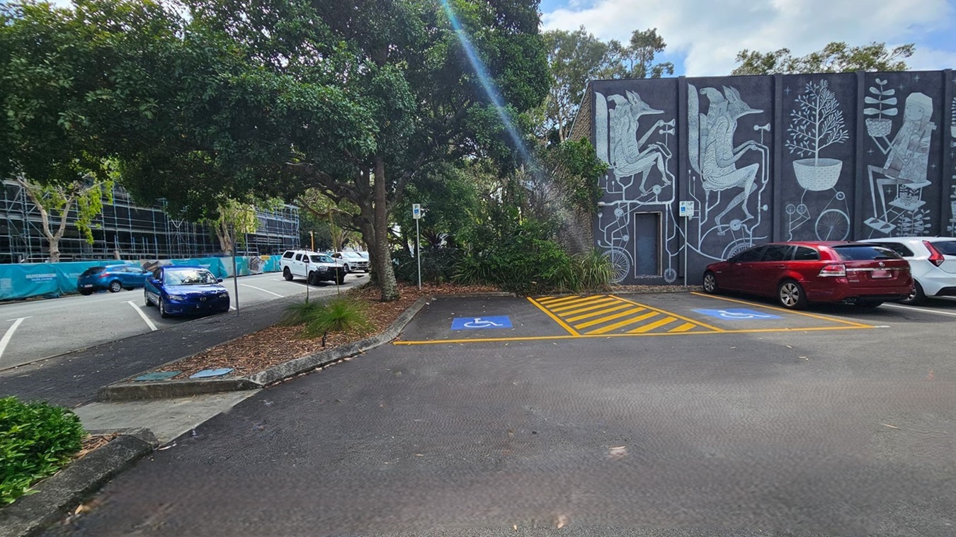 A car park with designated disabled parking spaces marked in blue and yellow on the right. A mural with abstract figures and plants is painted on the building wall in the background. On the left, a row of parked cars, including a blue car, is partially shaded by large trees. A construction site with scaffolding and a blue fence is visible further left. The ground is covered with scattered leaves.