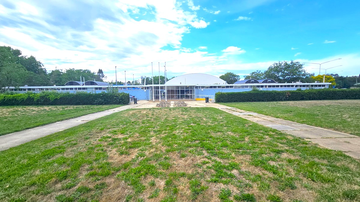 In the centre stands a concrete building with a dome-shaped roof. Trimmed shrubs border the front of the building. Adjacent to it lies expansive ticketed grassy parkland. Two concrete pathways lead to the building's entrance, and a modern concrete bike rack is positioned towards the end of the car park. Multiple street lights and trees surround the area.
