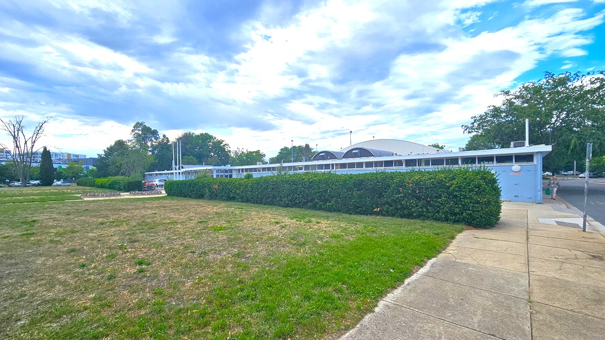 To the left, an expansive grassy parkland extends. On the right, a brick pathway and a road with a step-free entrance are present. Behind stands a blue concrete building with a dome-shaped roof. Trimmed ornamental shrubs border the building's front. Towards the far left, a concrete bike rack is positioned. The area is surrounded by trees.