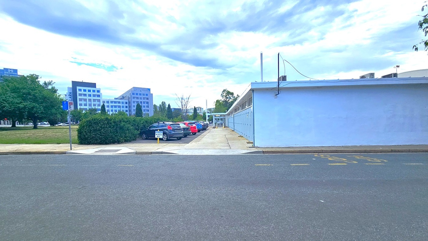 To the left lies an expansive grassy parkland, while on the right, a brick pathway and a road with a step-free entrance are present. Adjacent to the left is a car park with cars of various models and colors. A blue concrete building stands nearby, bordered by neatly trimmed ornamental shrubs. The surroundings include trees and commercial buildings.