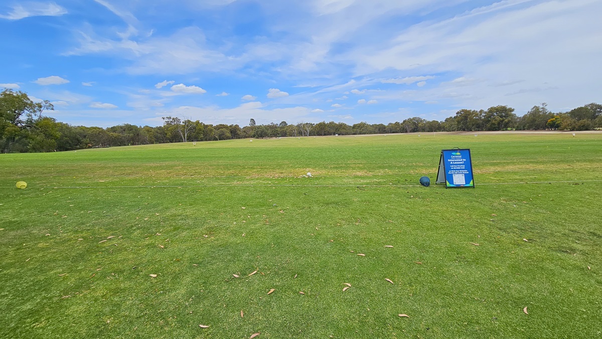 A concrete path directs towards the green. On the right, there is an elevated grassy area with a green sign reading "10th Tee." A golfer is visible near the freestanding banner. On the left, there is manicured grass. Further ahead, more trees are present.