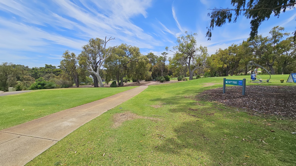 A concrete path directs towards the green. On the right, there is an elevated grassy area with a green sign reading "10th Tee." A golfer is visible near the freestanding banner. On the left, there is manicured grass. Further ahead, more trees are present.