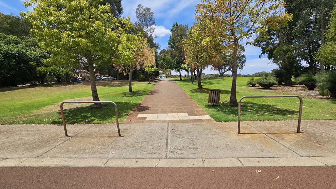 The car park is on a sloping hillside with a pink concrete floor. Three cars of various models and colors are parked under gum trees. A curving concrete path on the right ascends to an elevated area, bordered by tall trees. An accessible parking space, marked with a blue wheelchair symbol, is also visible.