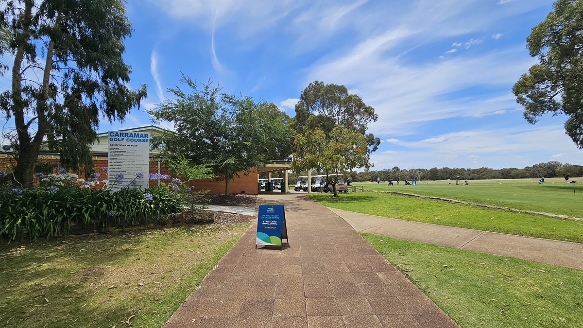 On the left stands a red brick building with a veranda. A large banner displaying facility information is positioned on the left. Flowering plants and tall trees border the left side. A blue free standing banner is placed in the middle of the brick pathway. To its right, a path descends to the green, dominating the spacious area. Golf carts are parked near the veranda.