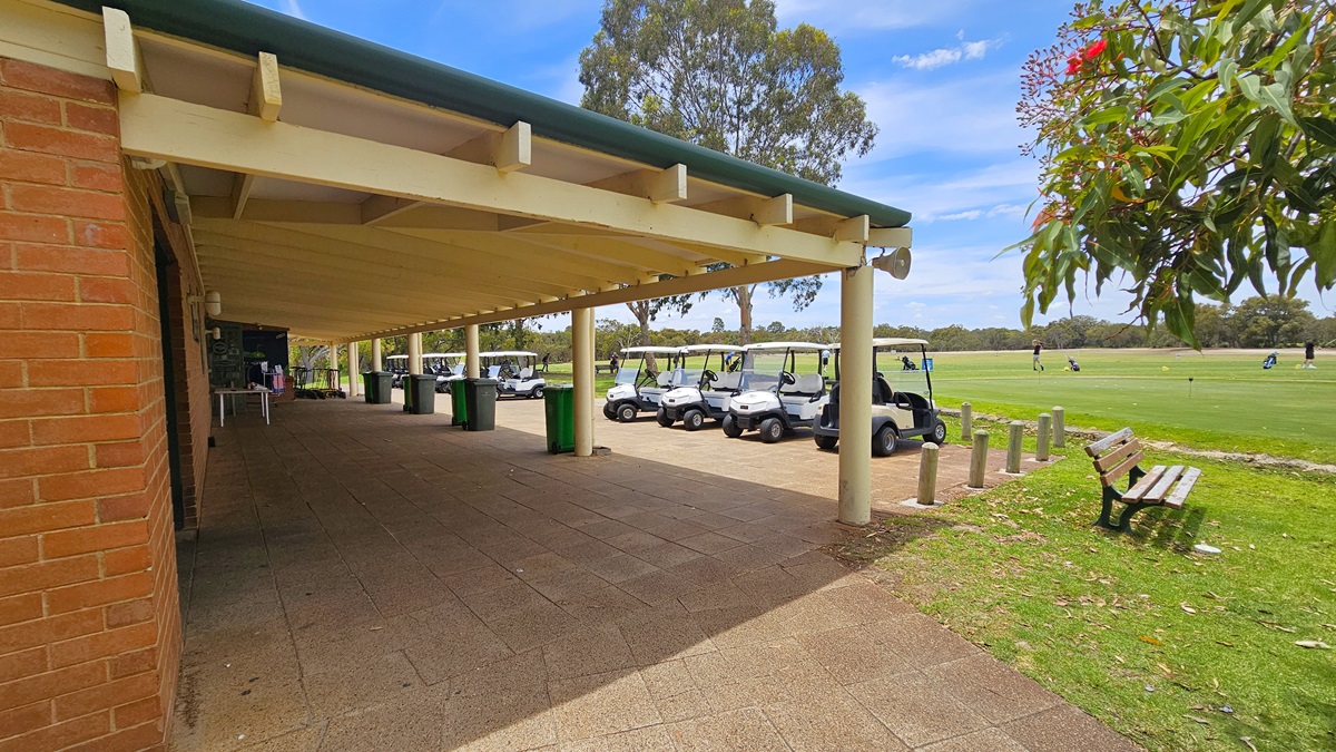 The veranda, constructed with corrugated roofing and exposed wooden beams, has adjacent parked golf carts. Large green rubbish bins, including a white table, are lined in front. The building's facade is made of red bricks. To the right, bench chairs are positioned near concrete bollards. The area on the right features a spacious green where a few golf players are visible.