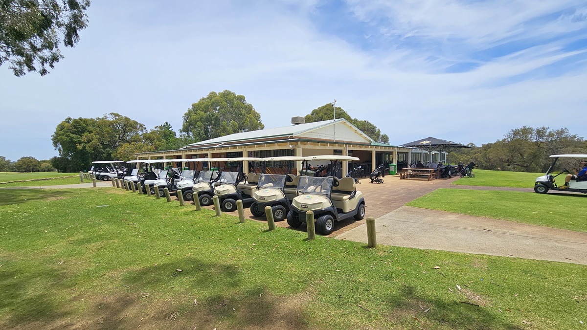 Golf carts are arranged in a row near the sloping grassy area, bordered by concrete bollards. Behind them, a low-rise building with a veranda is equipped with picnic tables under umbrella shades. On the right, a golf cart is parked. The surroundings feature green grass and tall trees.