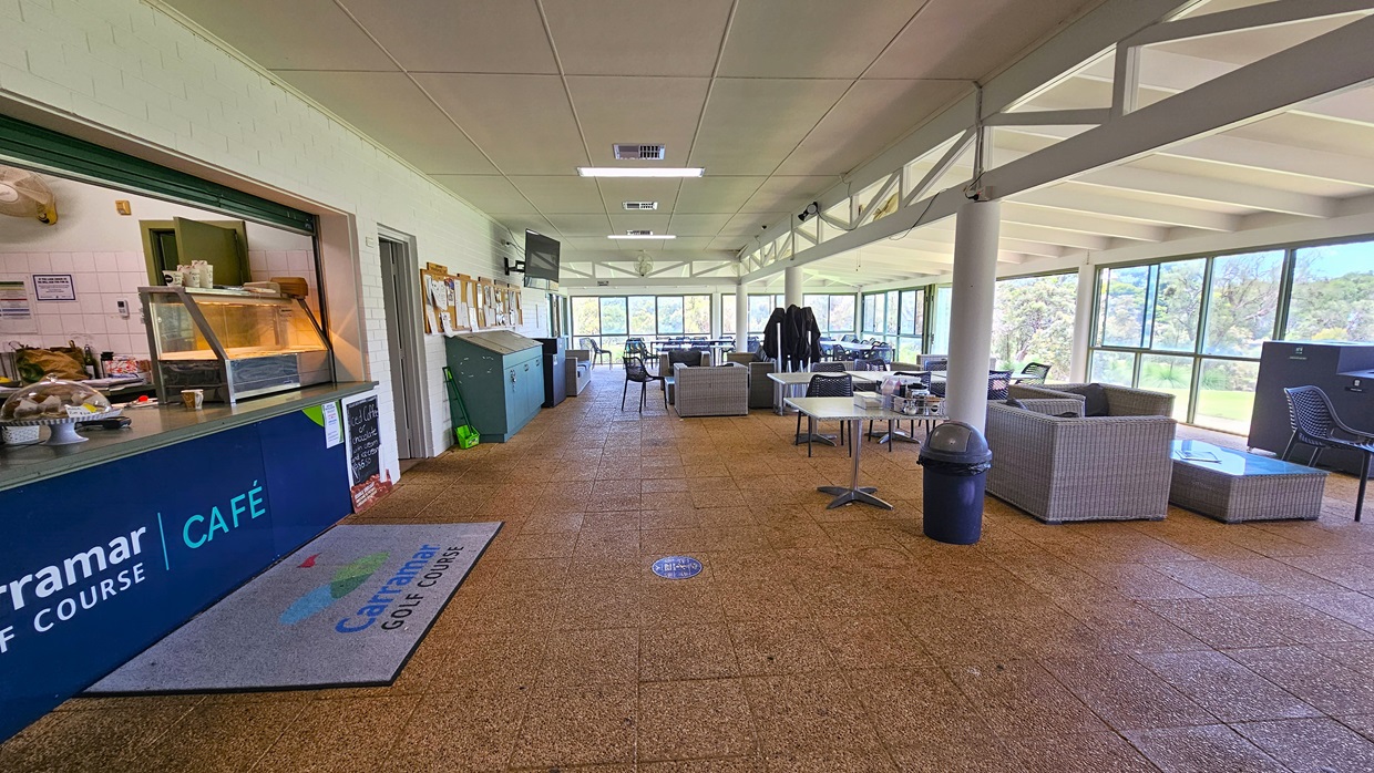 On the left, the cafe counter exhibits the facility's signage with a food rack. Adjacent is a corkboard for reminders, and cleaning accessories are underneath. To the right, there are cane-style couches and movable chairs and tables. A black rubbish bin is positioned near the middle post. The wall is made of white bricks, the ceiling is white, and the floor has brown tiles, featuring a large floor mat with the facility's name.