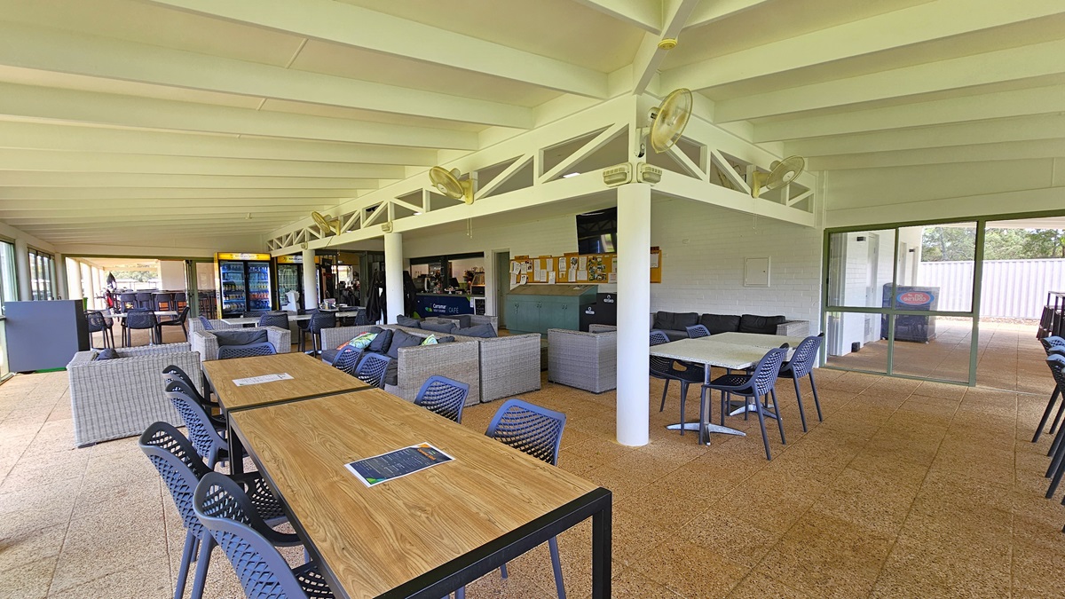 A glass door opens to a wide, spacious green area with manicured grass. Trees and shrubs surround the area, extending through the green. Concrete bricks fill the floor near the cafe.