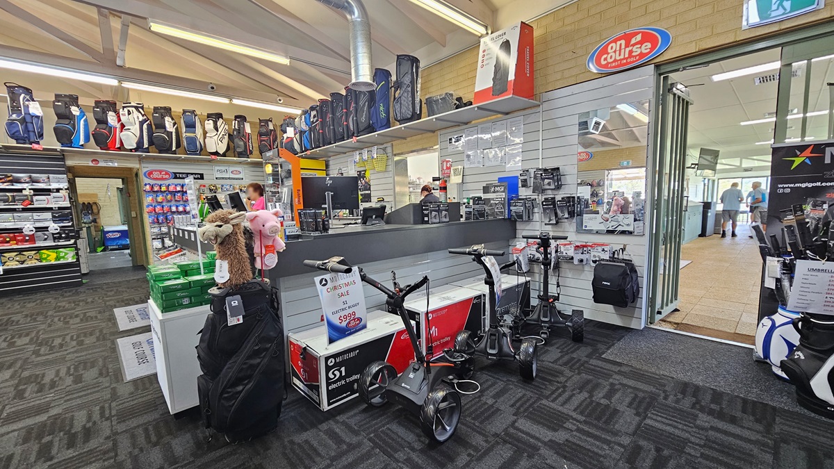 The reception counter, centrally positioned, accommodates three computers and retail items. Golf balls and bags are displayed on the left, while electric trolleys are showcased on the right. A glass door on the right opens outward, leading to an area with cream tiles. The ceiling is white with exposed metal beams and fluorescent lights, and the floor is covered with grey carpet.