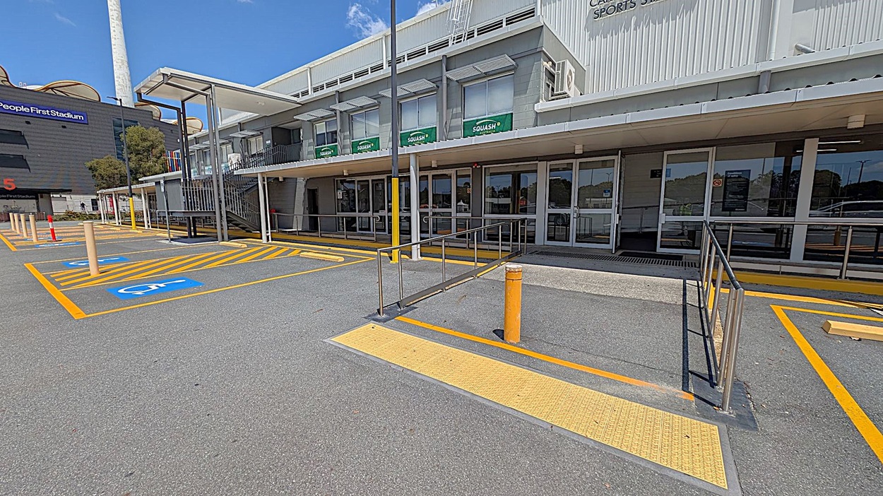 A parking area with designated disabled parking spaces marked in blue and yellow lines on the left. A ramp with metal railings leads to the entrance of a building on the right, which has large glass doors and windows with "SQUASH" signs. The building is grey with a flat roof and a covered walkway. A tall structure with a chimney is visible in the background on the left. The ground is paved with asphalt.