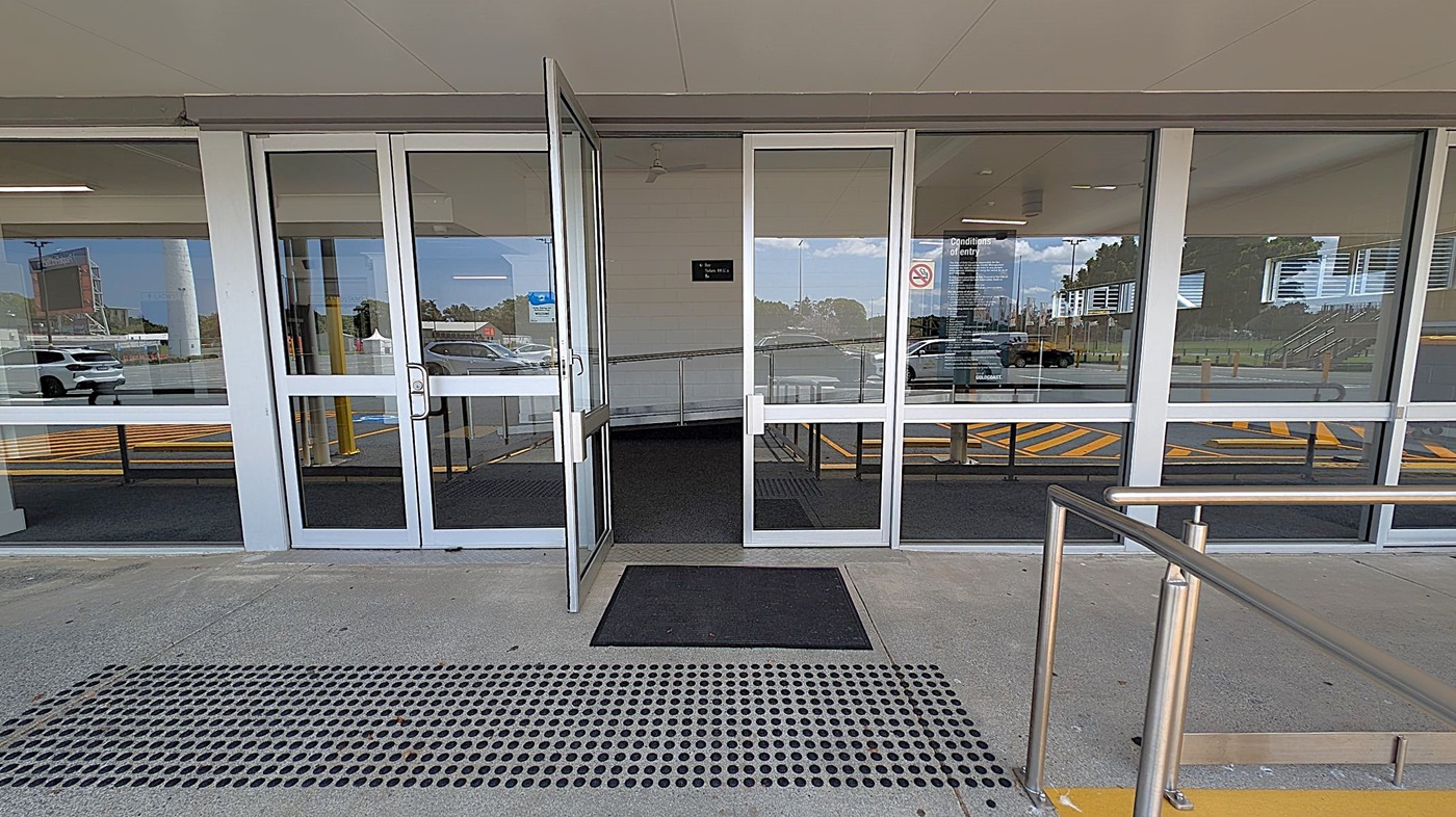 Glass entrance doors with metal frames, one door is open. A black mat is on the ground in front of the entrance, with a textured metal grate nearby. Inside, a ramp with metal railings is visible. Reflections of parked cars and a building are seen in the glass. Yellow lines are painted on the pavement outside. The ceiling is smooth and light-coloured.