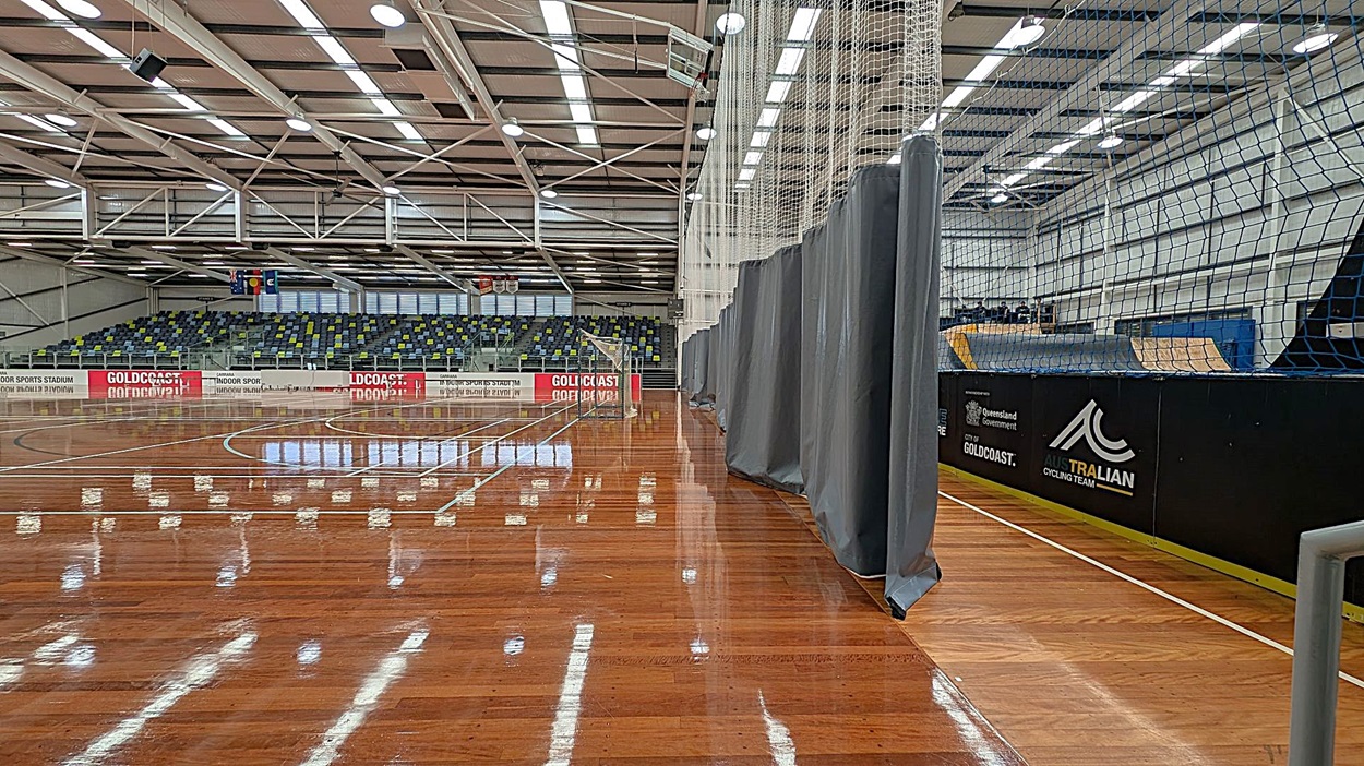 A large indoor sports hall with a polished wooden floor. On the left, there's a white wall with a fire exit door and a green "Push bar to open" sign. Above the door is an emergency exit light. A fire hose reel is mounted on the wall. In the middle, there are tiered seating with multicoloured chairs. The ceiling is high with exposed beams and fluorescent lights. On the right, a grey curtain partially covers the view of the court.
