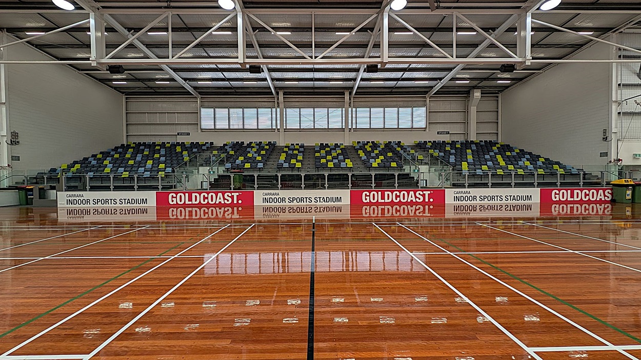A spacious indoor sports stadium with a polished wooden floor marked with white and green lines. In the background, there are tiered seating areas with multicoloured seats in shades of grey, green, and blue. Above the seats, large windows allow natural light to enter. The ceiling features exposed beams and lighting fixtures. Signs on the barrier in front of the seating read "Carrara Indoor Sports Stadium" and "Gold Coast."