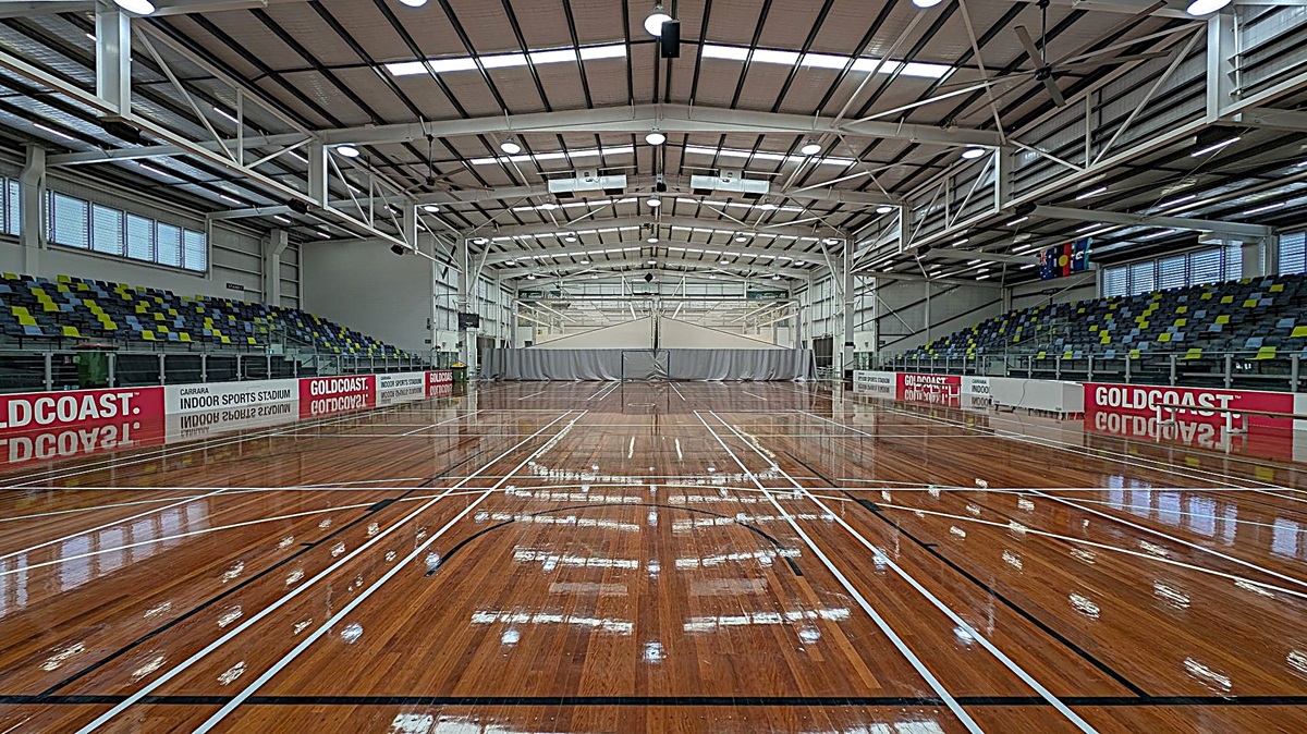 A spacious indoor sports stadium with a polished wooden floor marked for various sports. On the left and right are tiered seating areas with green, blue, and yellow seats. The walls have large windows and banners reading "GOLDCOAST" and "INDOOR SPORTS STADIUM." The ceiling is high with exposed beams and lighting fixtures. In the centre, a grey curtain divides the space.