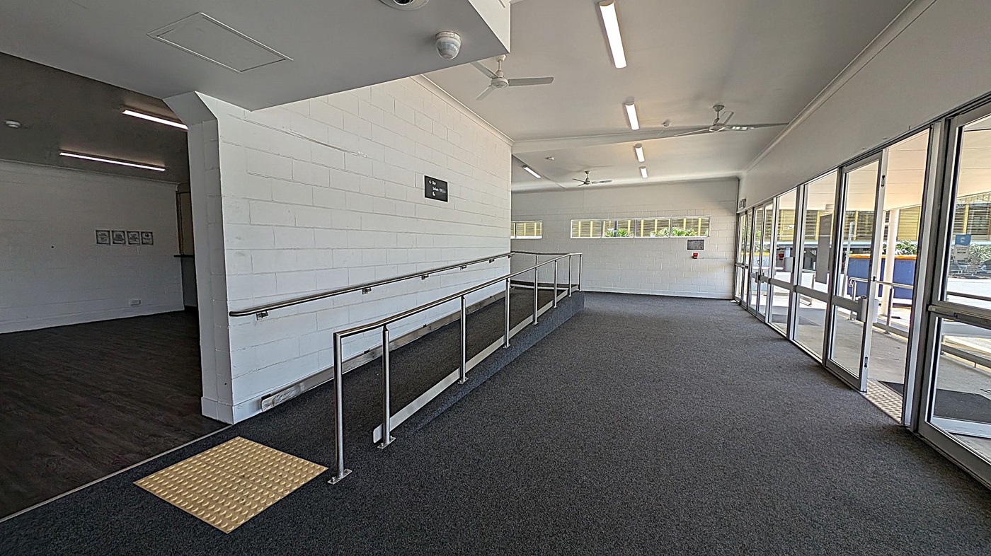 A corridor with a ramp on the left, featuring a metal handrail and a yellow tactile paving at the base. The floor is carpeted in dark grey. On the right, there are large glass doors and windows allowing natural light to enter. The ceiling is white with fluorescent lights and ceiling fans. The walls are painted white, and there is a small sign on the left wall. In the background, there are more windows and a view of an outdoor area.
