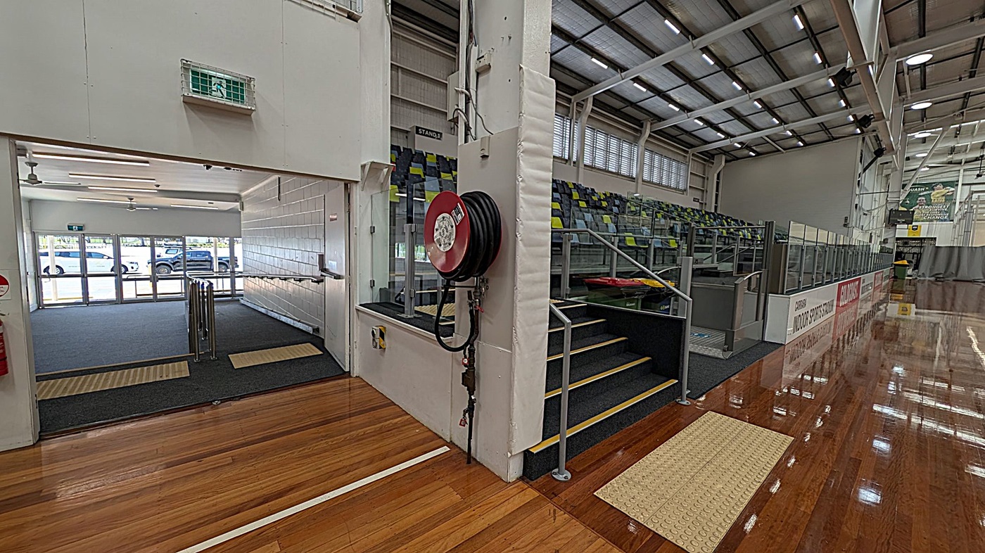 On the left, there's an open entrance with glass doors and a view of parked cars outside. A green exit sign is above the doorway. In the centre, a fire hose reel is mounted on a white pillar. To the right, there are tiered seating with green and yellow chairs, and a set of stairs with a handrail. The floor is polished wood, and the ceiling has exposed beams and lights.