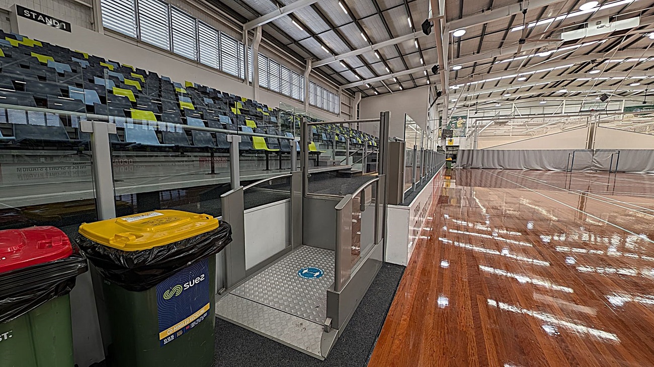 Indoor sports hall with a polished wooden floor on the right. On the left, there are tiered seating with alternating blue and green seats. In the foreground, there are three bins with red, yellow, and green lids. A ramp with metal railings leads up to the seating area. The ceiling is high with exposed beams and multiple lights. Large windows with horizontal slats are above the seating area.
