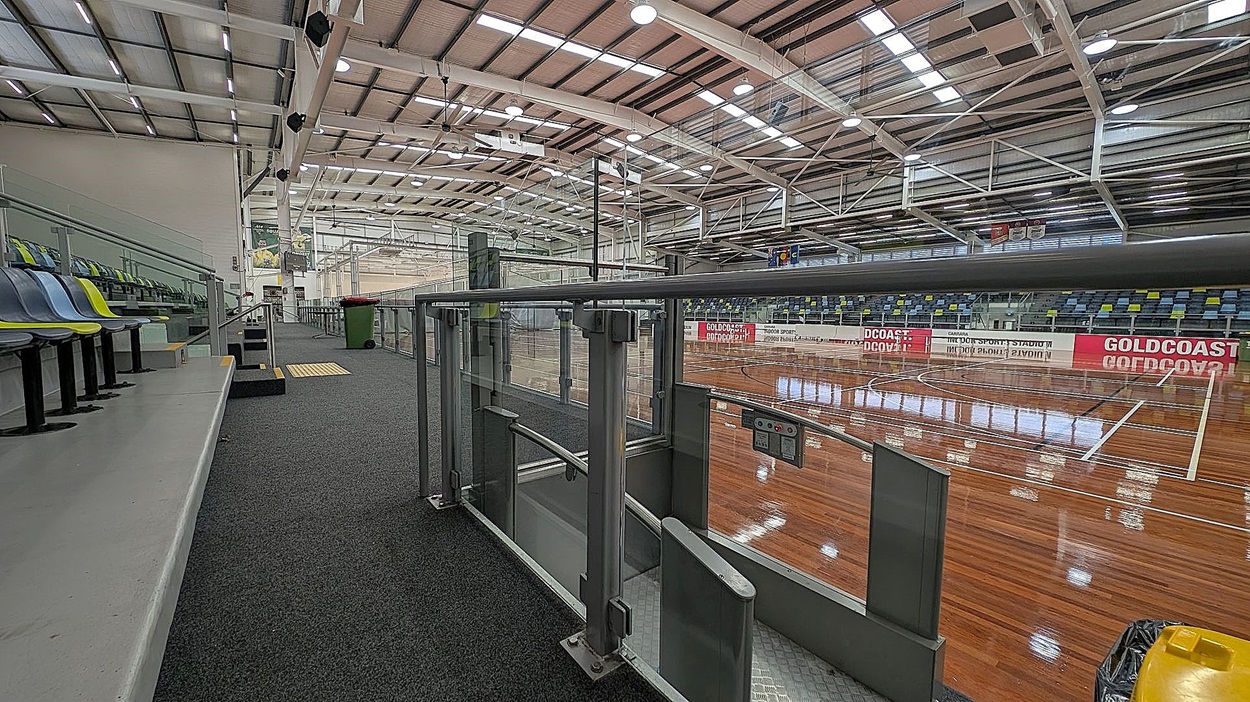 Indoor sports stadium with a polished wooden floor and marked courts. On the left, there are tiered seating with blue and green chairs. A grey carpeted walkway runs alongside the seating. In the foreground, a metal barrier with a gate is visible. The ceiling is high with exposed beams and bright lighting. On the right, a red and white sign reads "GOLDCOAST."