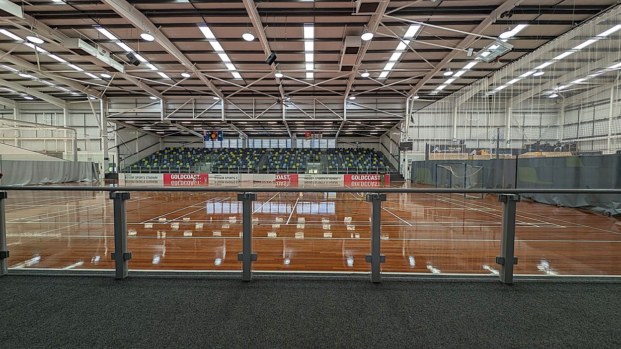 A large indoor sports stadium with a polished wooden floor and multiple marked courts. In the background, there are tiered seating areas with multicoloured seats. The ceiling is high with exposed beams and bright overhead lighting. A glass railing runs along the foreground, separating the viewing area from the courts. Banners and nets are visible on the right side, and the walls are lined with grey curtains.