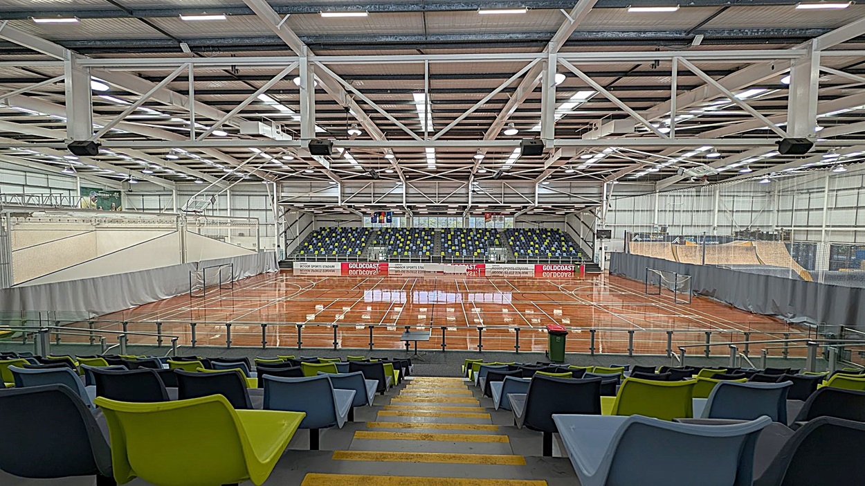 A large indoor sports arena with a polished wooden floor marked for various sports. In the foreground, there are rows of multicoloured plastic seats in blue, green, and black. The background features a tiered seating area with more multicoloured seats. The ceiling is high with exposed beams and bright lighting. Nets hang along the sides of the court, and banners are visible on the far wall.