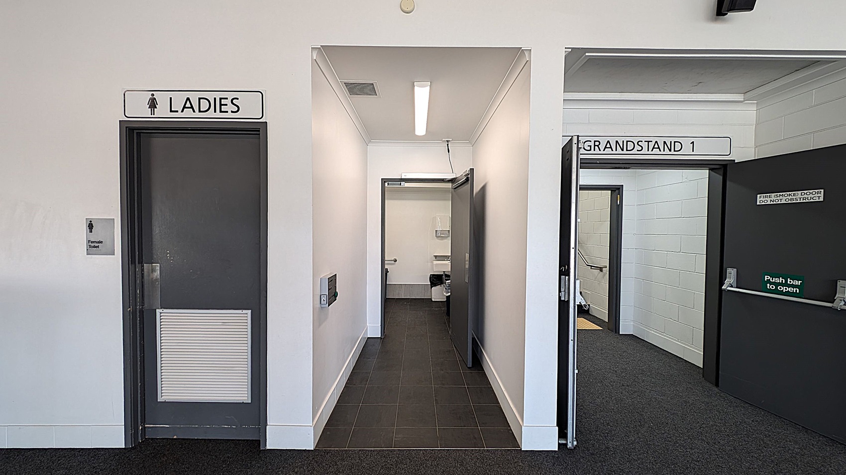 A spacious indoor area with a grey carpeted floor. On the left, there are open doors leading to a restroom and an entrance marked "Grandstand 1". In the centre, a bar area with a dark floor and a counter. On the right, large glass doors and windows allow natural light in, with a view of the outside. The ceiling is white with recessed lighting.