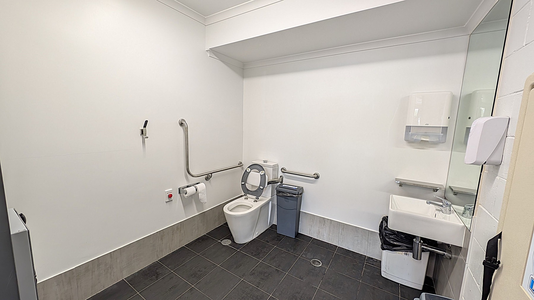 A clean, accessible bathroom with white walls and a dark tiled floor. On the left, there are grab bars and a toilet with a raised seat. A toilet roll holder and an emergency button are nearby. In the centre, a bin is placed next to the toilet. On the right, a sink with a mirror above it is mounted on the wall. A soap dispenser and a paper towel dispenser are also present. The ceiling is plain white with a small recessed area.