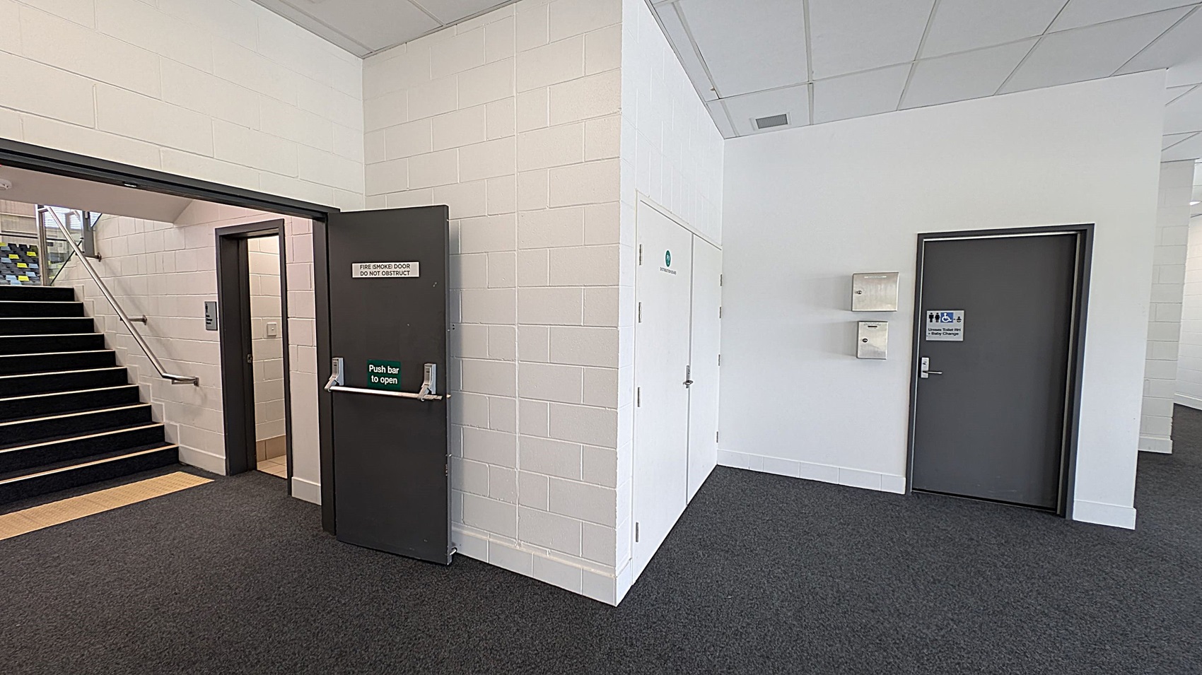 A corridor with a dark grey fire door on the left, labelled "Fire Smoke Door Do Not Obstruct," leading to a staircase with a metal handrail. The floor is carpeted in dark grey. On the right, there is a grey door with a sign indicating accessible toilets. Above this door are two wall-mounted boxes. The walls are painted white, and the ceiling is made of white tiles.