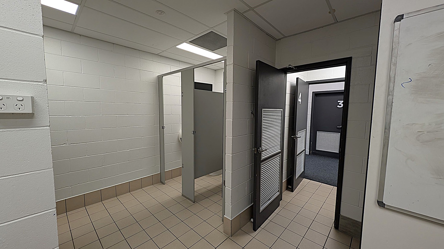 A corridor with tiled flooring and white brick walls. On the left, there are two open cubicles with grey doors. On the right, two closed doors numbered 3 and 4, each with a vent panel. The ceiling is fitted with fluorescent lights. A whiteboard is partially visible on the far right wall.