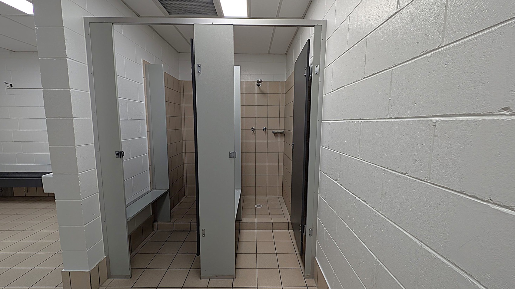 A public shower area with beige tiled walls and floor. Two showerheads and a small shelf are visible in the middle. The shower is enclosed by grey partitions with a door slightly ajar. The surrounding walls are white-painted brick. To the left, a glimpse of a green-tiled floor and a bench can be seen. The ceiling is white with fluorescent lighting.