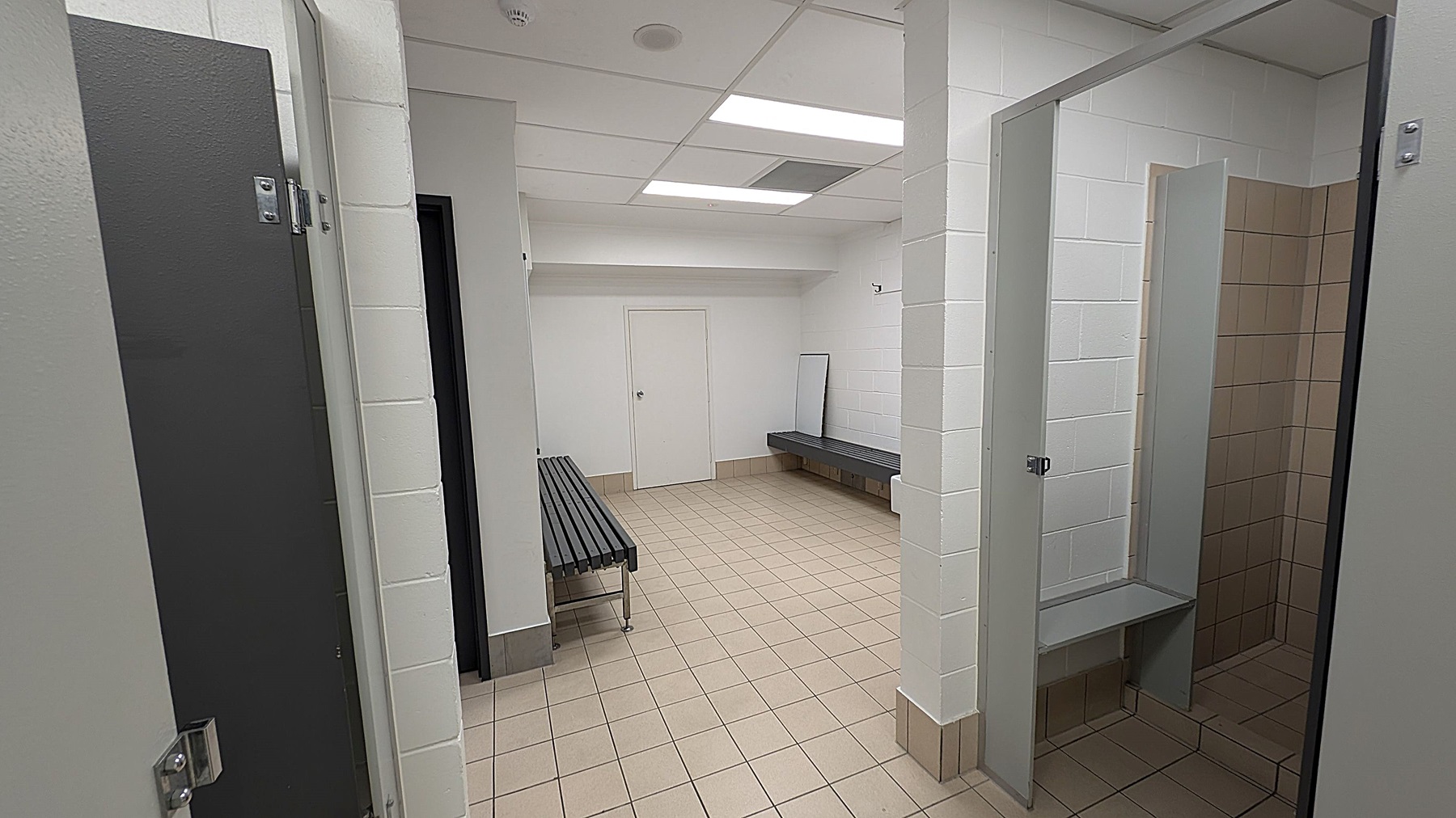 A locker room with beige tiled flooring and white tiled walls. On the left, there is a dark grey door. In the centre, a long grey bench is positioned against the wall, with a mirror leaning beside it. On the right, there is an open shower stall with beige tiles. The ceiling is white with recessed lighting.