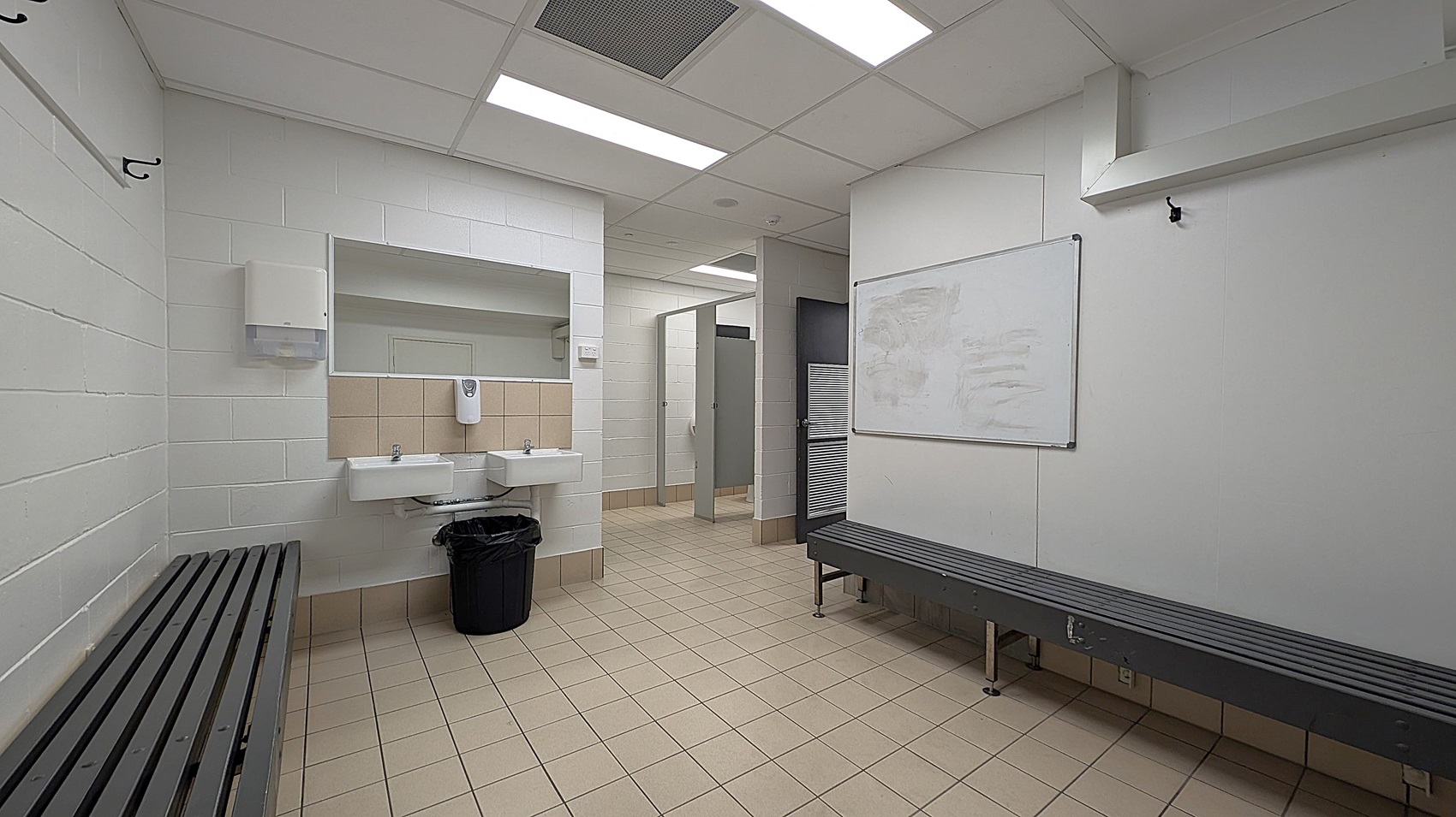 A locker room with beige tiled flooring and a white tiled ceiling with fluorescent lights. On the left, there is a black bench and wall hooks. In the centre, two white sinks with a soap dispenser and a black bin are mounted on a tiled wall. In the background, there are open doorways leading to shower or toilet stalls. On the right, another black bench and a whiteboard are visible.
