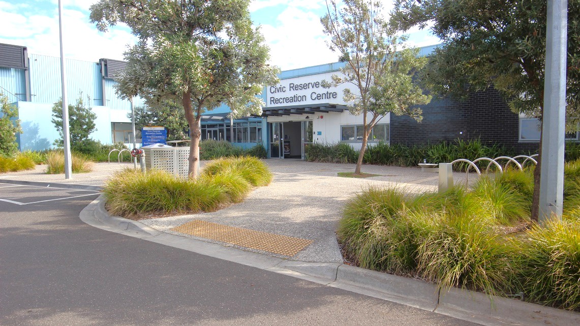 The foreground features a paved area with light-coloured stones and scattered grass patches. On the left, there’s a grey parking space marked with a single parking sign. The entrance of the Civic Reserve Recreation Centre is centrally positioned, featuring large windows and a prominent sign above the door. To the right, there's a landscaped area with shrubs and small trees, along with a decorative garden edging.