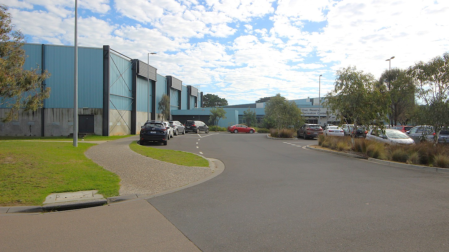 In the foreground, a curved asphalt road leads past landscaped grass areas on the left, bordered by a gravel path. Several vehicles are parked along the right side of the road, with one black car prominently displayed. To the left, the building features a blue exterior with vertical paneling, and there are light poles positioned throughout the area. In the background, the Civic Reserve Recreation Centre is visible, identified by its signage, with more vehicles parked in a lot beyond.