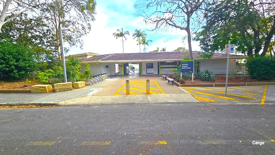 Front of Cleveland Aquatic Centre. To the right are accessible parking spaces marked by yellow diagonal lines, a mobility disability logo parking sign, and a no-stopping sign. Behind it is a large blue facility name signboard and two low wooden benches. In the middle of the entryway are yellow diagonal lines enclosed within a rectangle and yellow bollards. To the left are three large yellow rectangular rocks and bike racks. To the back are low-set cream buildings with brown tiled roofing.